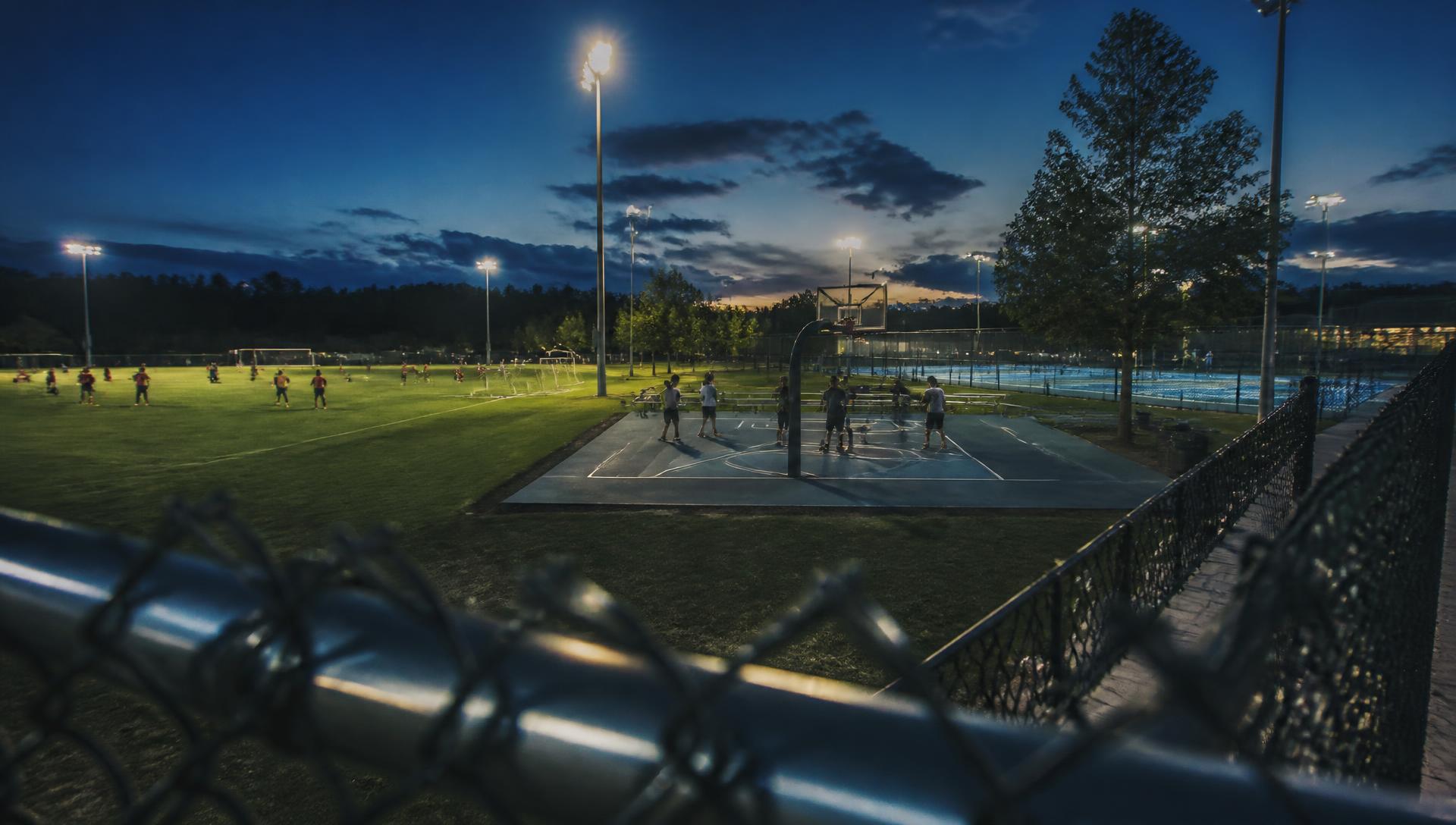 Floodlit multi-sport training complex at dusk with football, basketball and tennis facilities in use